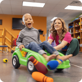 A Woman helping a child with needs and helping him play on a fun car.