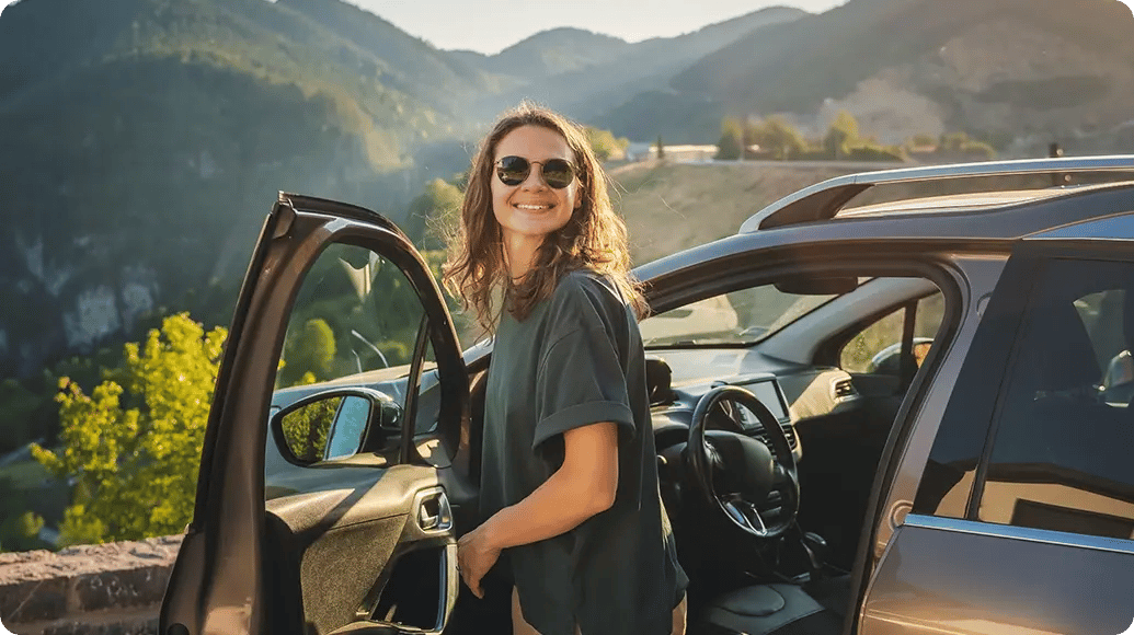 A person with shoulder-length hair stands beside an open car door, one hand resting inside the vehicle, wearing a casual short-sleeve top, with rolling green mountains and warm sunlight stretching across the landscape behind them.